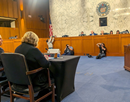 A congressional hearing room with attendees seated at a witness table facing a panel of lawmakers. Photographers are positioned in front of the dais, and the U.S. Senate seal is displayed on the marble wall behind the panel