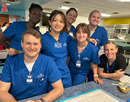 Group of nursing students and an instructor wearing blue scrubs pose together in a community health setting with tables and medical equipment visible in the background
