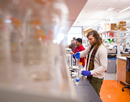 Person working at a laboratory bench, handling equipment or materials in a research setting. Shelves, glassware, and laboratory supplies are visible throughout the brightly lit lab, with additional people working in the background. The foreground is softly blurred, drawing focus to the researcher and the active laboratory environment