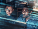 Two people looking at computer screens, an overlay of binary code in the foreground.
