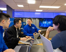 Faculty and students collaborate around laptops in a technology workspace, discussing projects in front of screens displaying UB AI branding