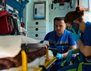 Two EMTs in an ambulance with a patient, one EMT uses a cell phone to communicate with the hospital