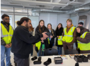 A group of students wearing high-visibility safety vests gathers around a table in a laboratory or classroom setting while an instructor demonstrates a handheld device. Equipment cases, electronics, and boxed materials are spread across the table as the students observe the demonstration. Exposed ductwork, glass partitions, and modern lab furnishings are visible throughout the brightly lit space