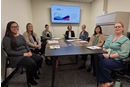 Seven people are seated around a conference table in a small meeting room. A wall-mounted screen displays information about a center focused on women's health, and notebooks and printed materials are arranged on the table