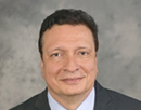 Professional headshot of a person wearing a dark suit and yellow tie against a gray studio background