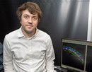 assistant professor Levi Todd seated next to a computer displaying a colorful scientific data visualization in a laboratory setting