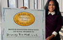 A person stands beside a large plaque mounted on an easel recognizing an award. The plaque features the text “Best Doctors in America” and “Rothschild” along with additional award information. The person is dressed in professional attire and is positioned next to the display in an indoor setting with large windows in the background