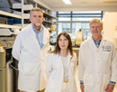 Three researchers wearing white laboratory coats stand together in a biomedical research laboratory. Scientific equipment, shelving, and gas cylinders are visible around them, highlighting an active research environment