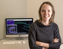 Researcher stands beside a computer displaying microscopy or imaging data in an office or laboratory workspace. The monitor shows colorful scientific images and analysis results