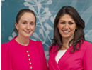 Two individuals stand side by side for a portrait against a blue backdrop featuring a decorative pattern. Both are wearing bright pink jackets, with one also wearing a pink top. The image is cropped from the chest up and has a professional, event-style appearance