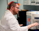 Man with rainbow mohawk in a lab coat in front of a high-throughput/plate-based cell imaging