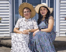 Two people wearing wide-brimmed sun hats sit together on the front steps of a house. Flowering plants in decorative pots line both sides of the entryway, and white siding and a blue front door frame the scene. The pair pose side by side outdoors in bright daylight, creating a warm portrait in a residential setting