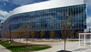 Modern multi-story research and innovation building with a curved glass façade and white metal exterior panels. Large floor-to-ceiling windows span the length of the structure, reflecting the sky and clouds. Landscaped walkways, young trees, and decorative lampposts surround the building, while a small glass pavilion stands in the foreground on a bright day