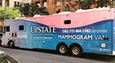 A large mobile mammography van from Upstate University Hospital is parked along a city street. The vehicle features a blue and pink exterior with prominent text promoting breast cancer screening services, including a phone number to schedule a mammogram. Buildings, trees, and parked vehicles line the street in the background